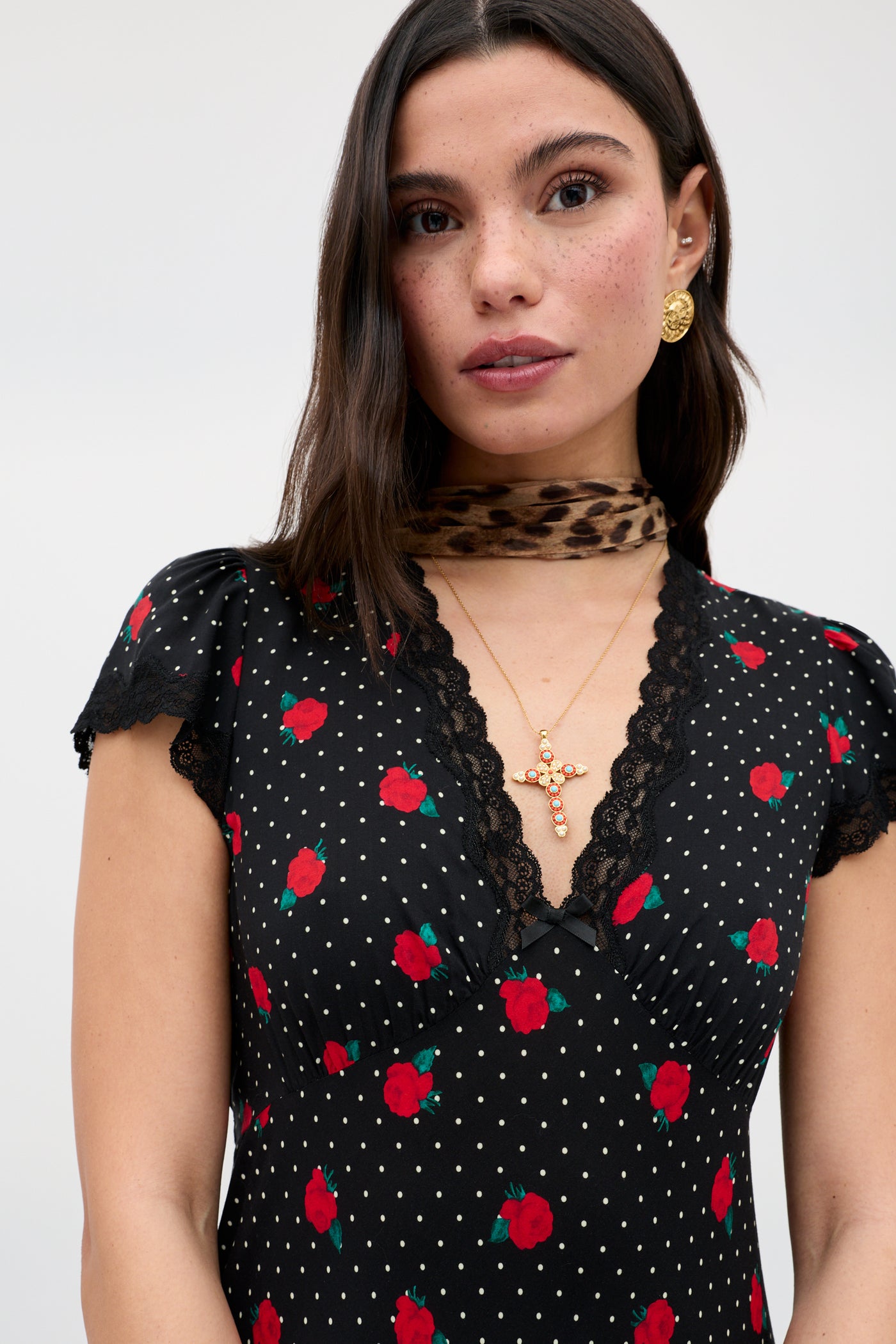 A woman with long dark hair and freckles wears the Clarice - Rose Spot Black dress, featuring red roses and white dots, paired with gold earrings, a gold cross necklace, a leopard-print scarf, and a lace collar. She has a neutral expression.