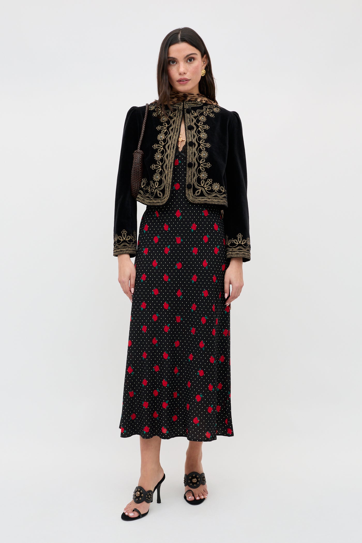 A woman wears the Clarice - Rose Spot Black dress, featuring red and white polka dots, paired with a black embroidered jacket, a small purse, and black heeled sandals. She stands facing forward against a plain white background.