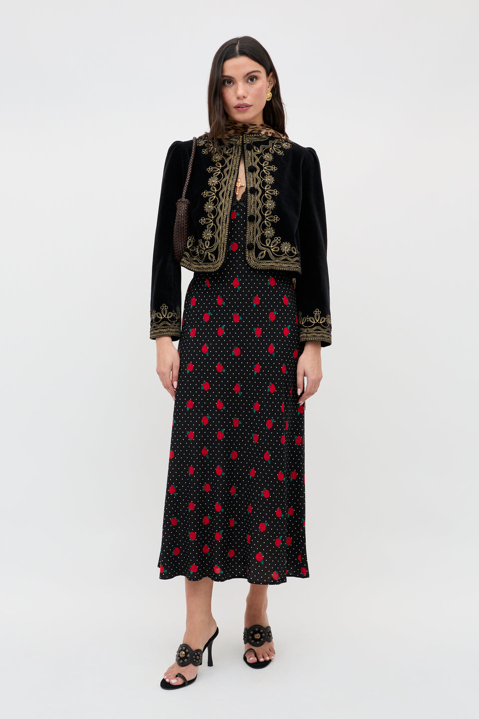 A woman wears the Clarice - Rose Spot Black dress, featuring red and white polka dots, paired with a black embroidered jacket, a small purse, and black heeled sandals. She stands facing forward against a plain white background.