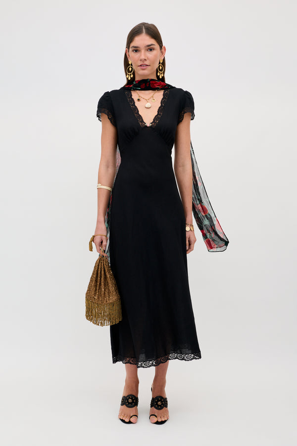 A woman wearing the Clarice - Black midi dress with lace trim, black sandals, gold statement earrings, a sheer floral scarf, and a fringed gold handbag stands against a light background.