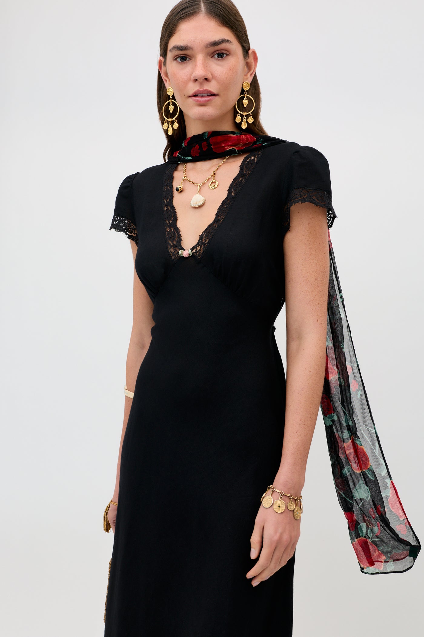 A woman wears the Clarice - Black midi dress with lace trim, layered gold jewelry, and a sheer red and green patterned scarf against a plain light background.