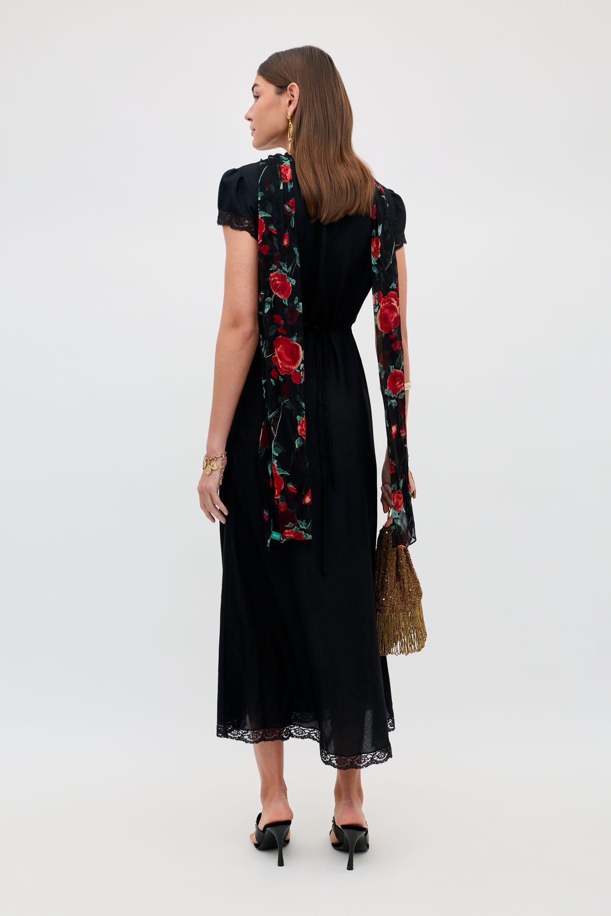 A woman stands facing away, wearing the Julien - Claudia Rose Black short-sleeve dress featuring a lace hem. She accessorizes with a long red floral hand-painted vintage scarf, gold fringed handbag, black heels, earrings, and a bracelet. Her hair is down, and the look is elevated by the fusion of timeless elegance and artistic detail offered by the scarf.