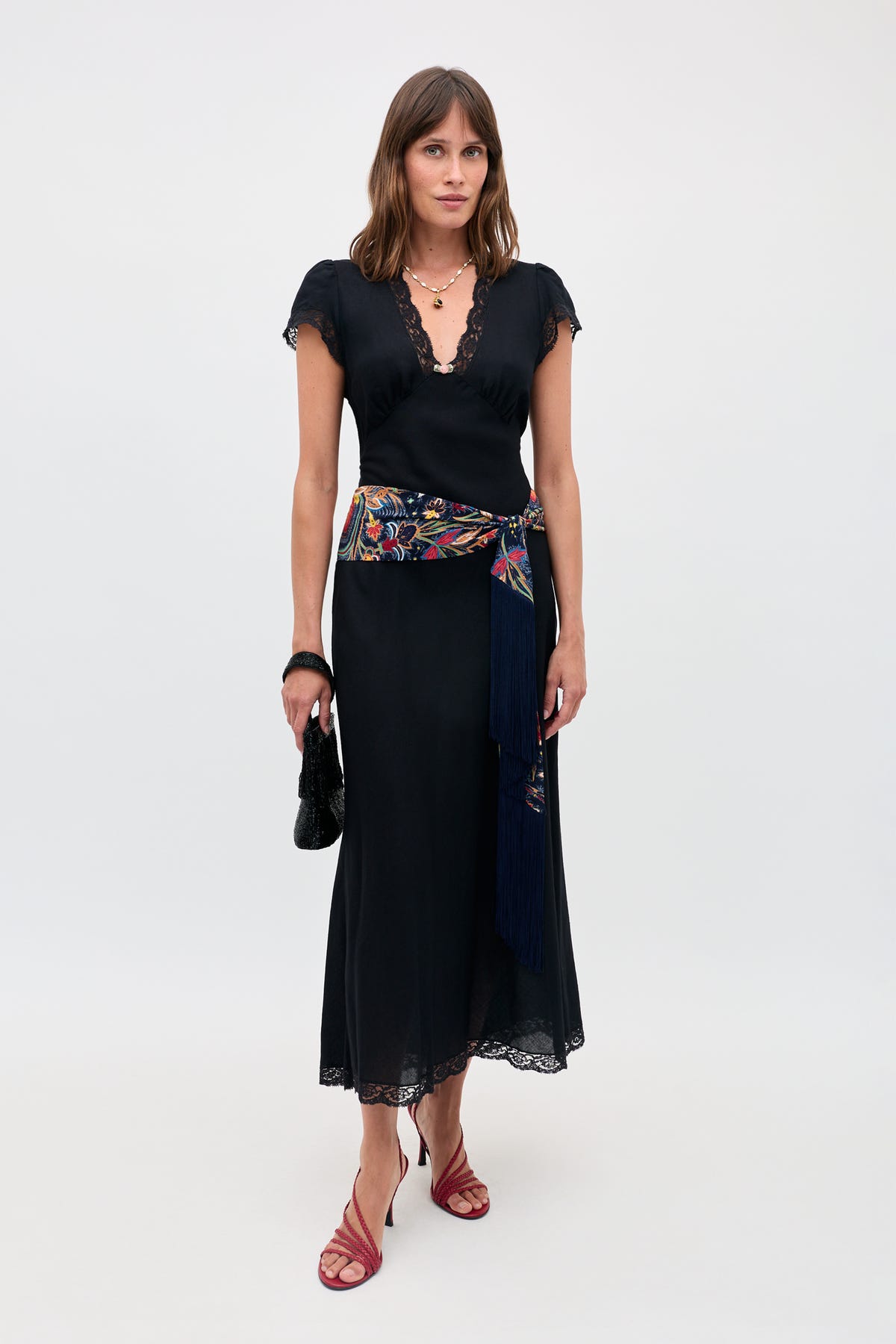 Wearing the Clarice - Black dress with lace trim, a vibrant embroidered sash with fringe, red strappy heels, layered necklaces, and holding a small black handbag, a woman stands against a plain light background.
