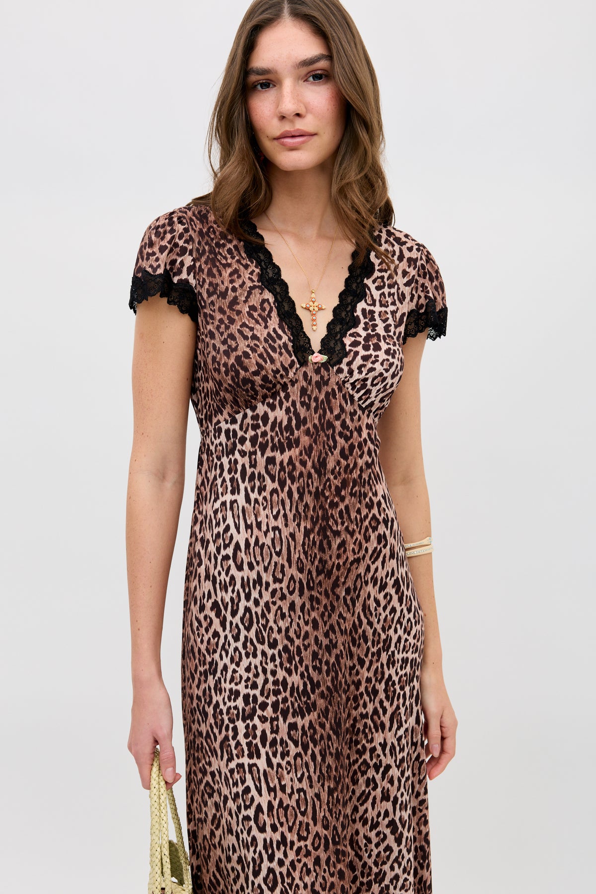 A woman stands against a plain background wearing the Clarice - Brown Leopard dress, featuring a leopard print with black lace trim on the neckline and sleeves. She holds a gold handbag and wears matching gold jewelry, looking directly at the camera.