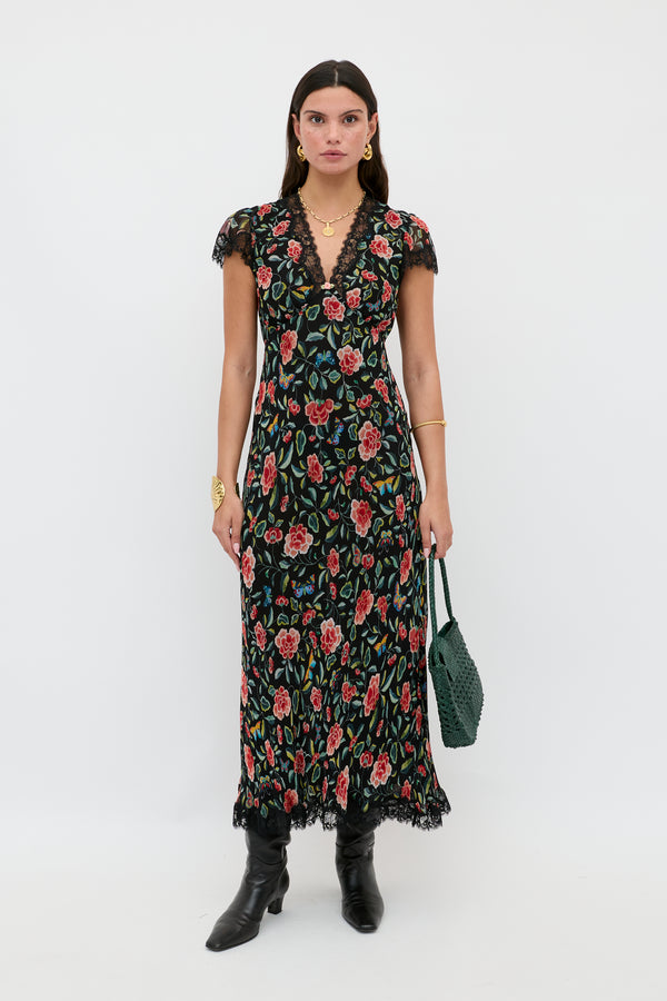 A woman stands against a white background in the Clarice - Butterfly Floral Black ankle-length crinkle chiffon dress with lace trim, styled with black boots, gold jewelry, a green woven bag, and straight dark hair.