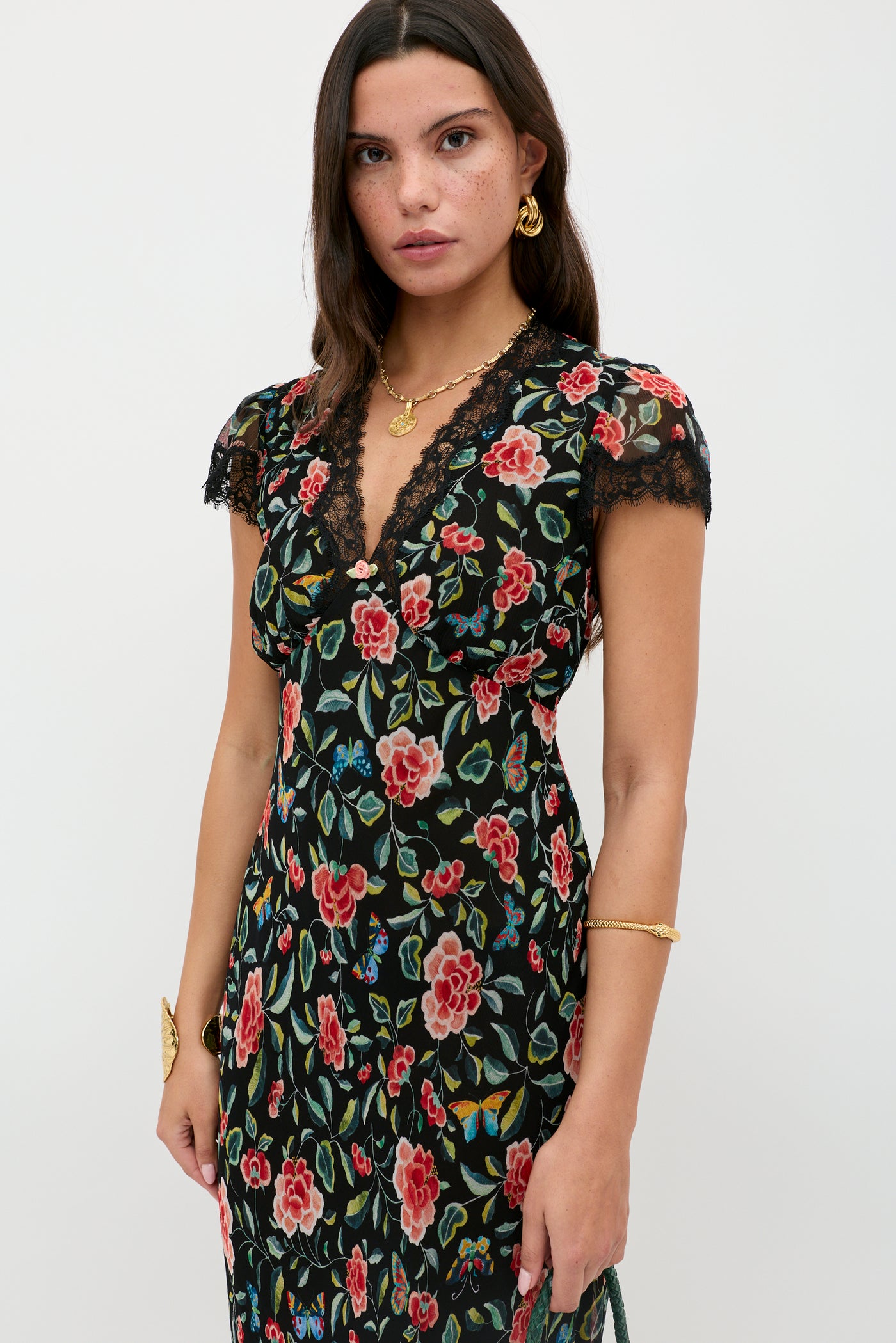 A woman with long dark hair wears the Clarice - Butterfly Floral Black dress, featuring a black crinkle chiffon fabric with colorful butterfly floral print and lace trim, accessorized with gold jewelry against a white background.