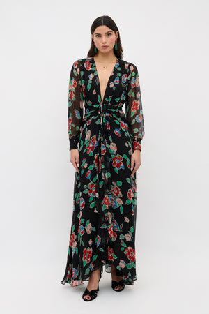 A woman with straight dark hair and a neutral expression stands against a plain white background, wearing the Meera - Kimmie Floral Charcoal long black dress with colorful floral print, sheer long sleeves, deep V-neckline, and black heeled sandals.