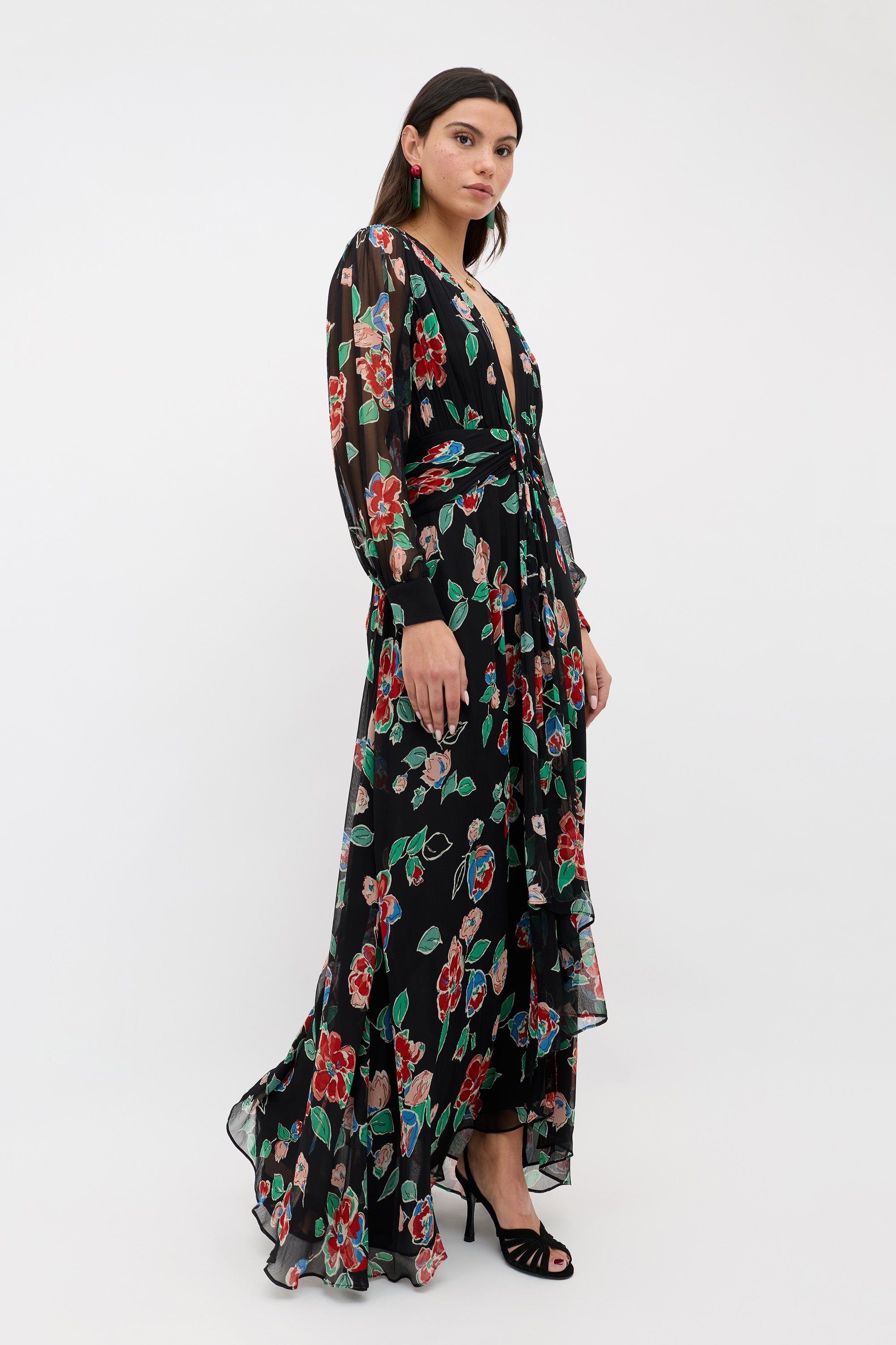 A woman in the Meera - Kimmie Floral Charcoal, a long sheer black dress with red and green floral patterns, stands in black high-heeled sandals against a plain white background.