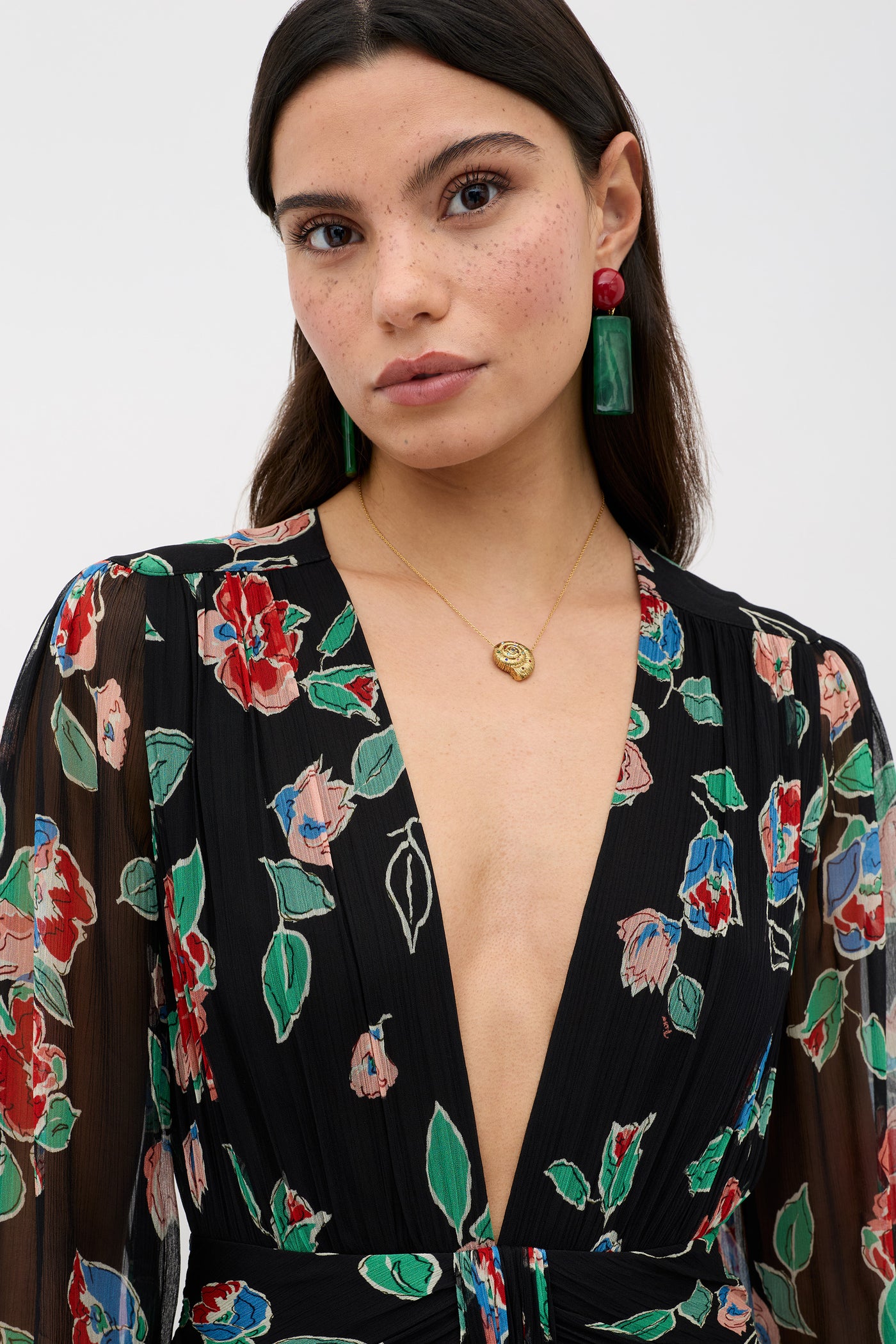 A woman with long dark hair wears the Meera - Kimmie Floral Charcoal dress with a deep V neckline, gold necklace, green rectangular earrings, and red circular earrings, standing against a plain light background.