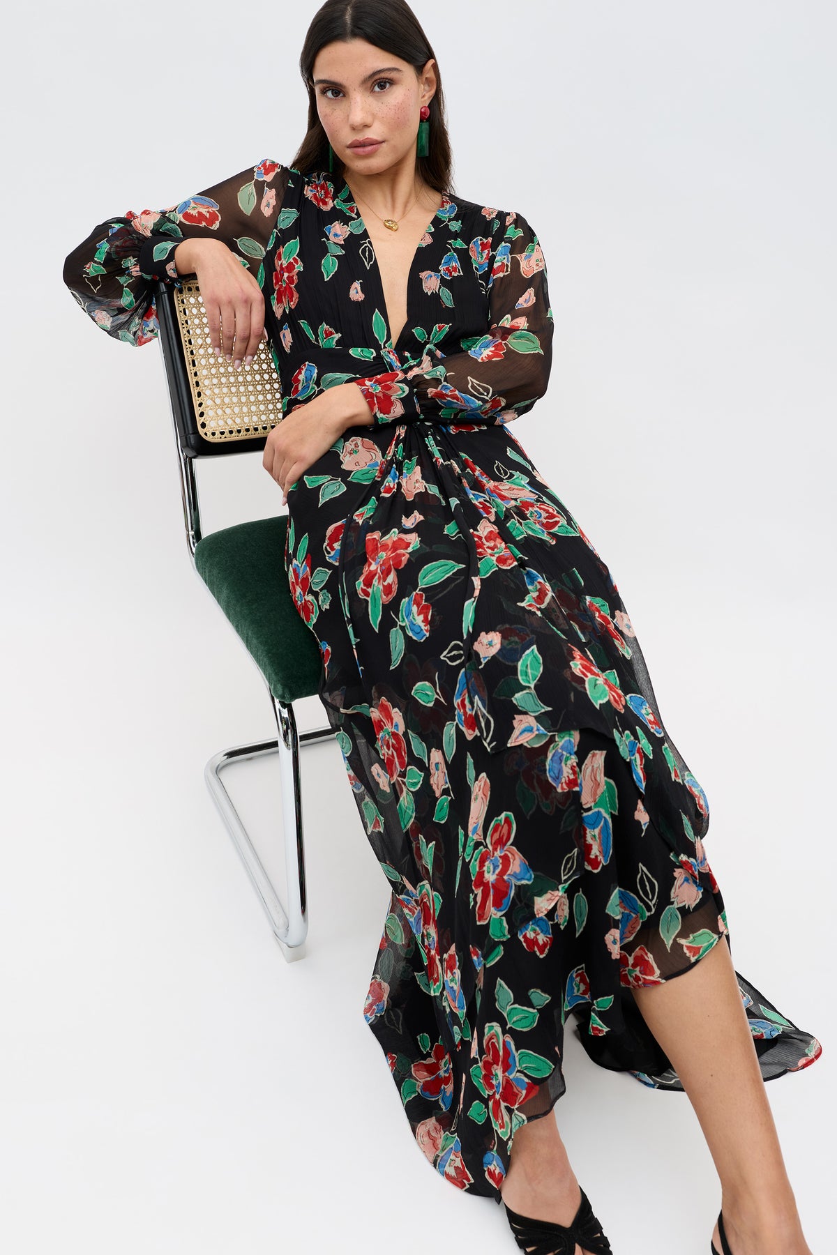 A woman with straight brown hair sits on a chair wearing the Meera - Kimmie Floral Charcoal dress, featuring a long black silhouette with colorful floral print and sheer sleeves. She pairs it with black open-toe heels and looks at the camera relaxed.