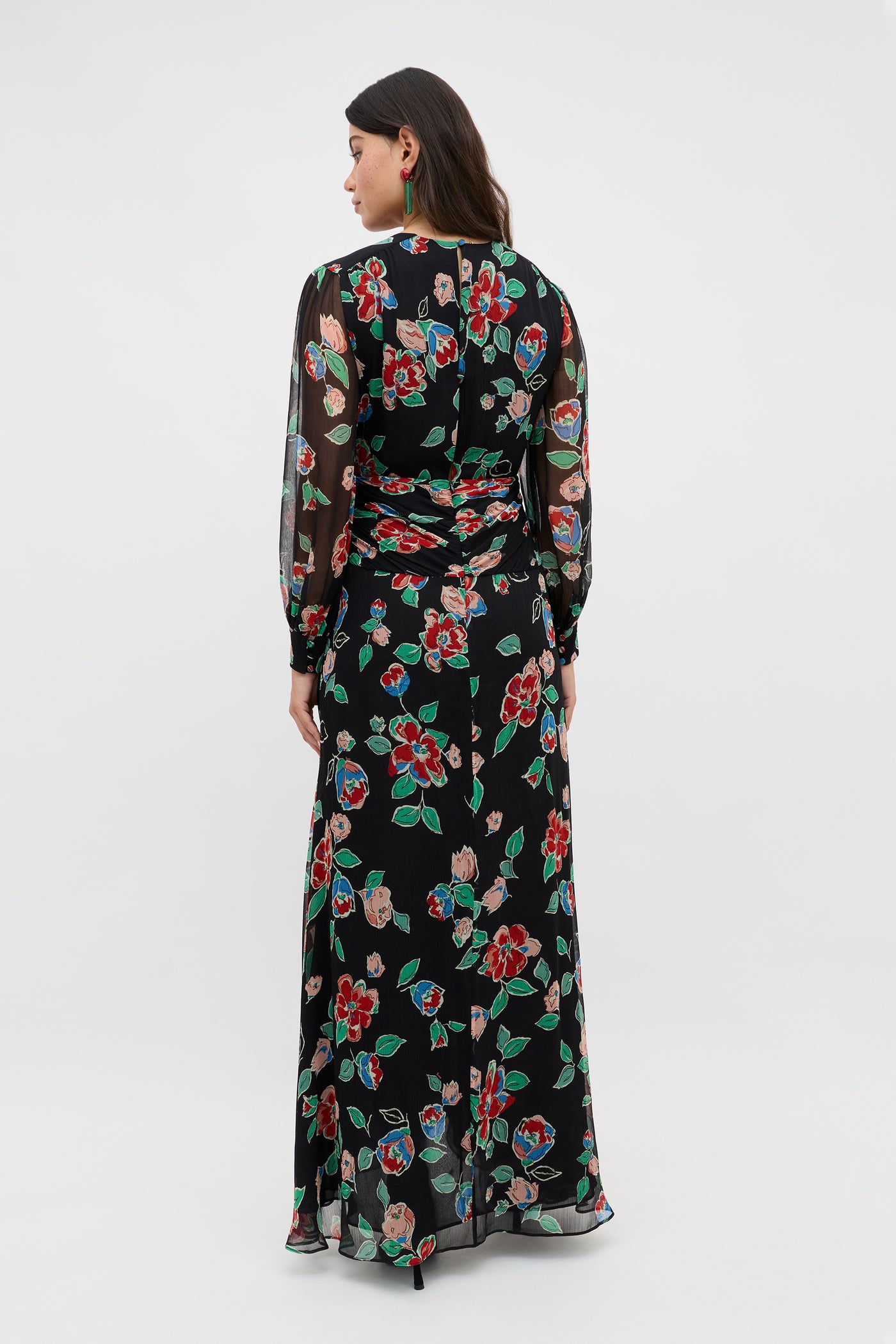 A woman faces away wearing the Meera - Kimmie Floral Charcoal dress, a long black sheer piece with long sleeves, a fitted waist, and vibrant red, green, and blue floral patterns.