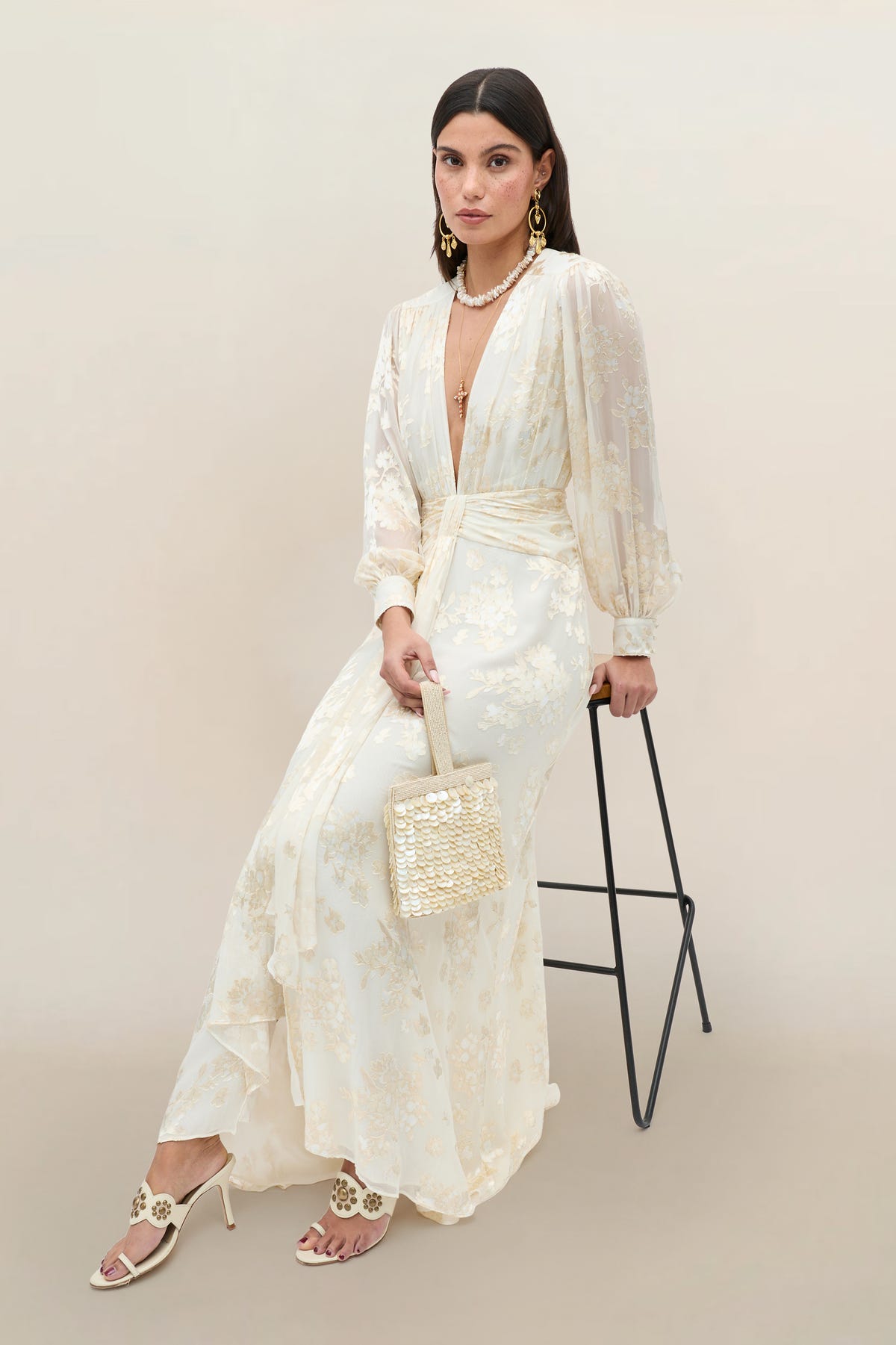 Wearing the Meera - Bouquet Devoré Cream silk-satin gown with subtle floral patterns, a woman poses elegantly on a stool in heeled sandals, statement earrings, and holding a small woven handbag against a neutral background.