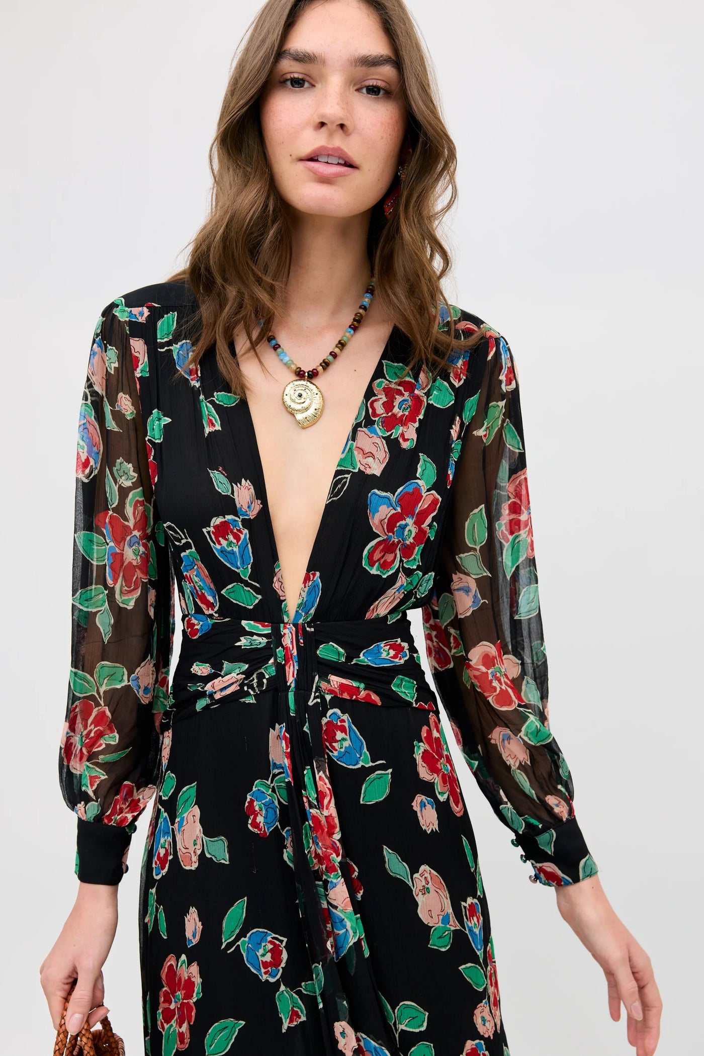 A woman wears the Meera - Kimmie Floral Charcoal dress featuring a colorful floral pattern, sheer long sleeves, and deep V-neckline. She has wavy brown hair and accessorizes with a beaded necklace and gold pendant.
