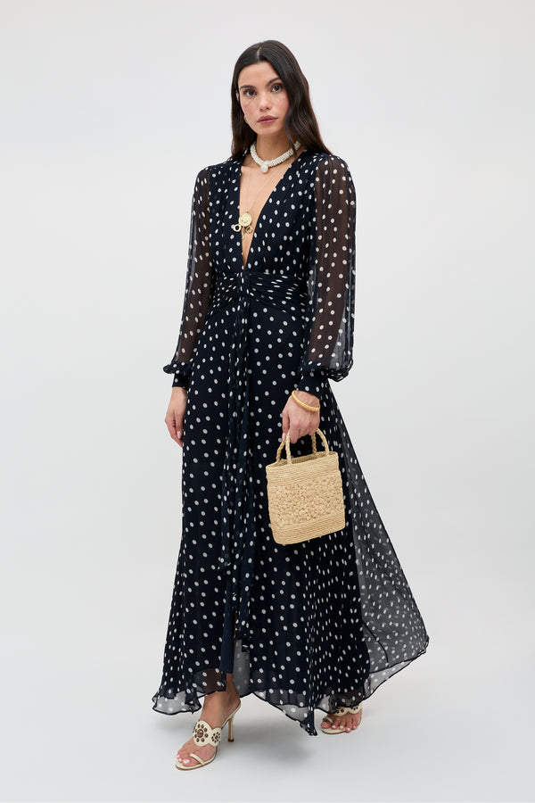 A woman wears the Meera - Bohemia Spot Deep Navy gown with long sleeves and a plunging neckline, paired with heeled sandals, layered necklaces, and a small straw handbag. She poses against a plain light background.