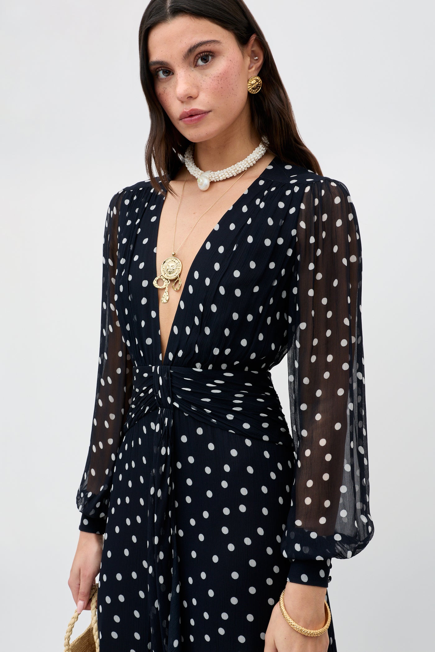 A woman wears the Meera - Bohemia Spot Deep Navy sheer long-sleeve polka-dot gown, styled with layered gold and pearl necklaces, gold earrings, and bangles, standing against a plain light background.