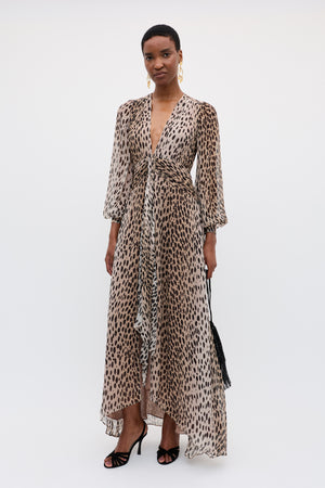 A woman wears the Meera - Antelope Spot Black gown with crinkled-chiffon fabric, leopard print, long sleeves, and a deep V-neckline. She pairs it with black heels, a small black bag, and gold earrings against a white background.
