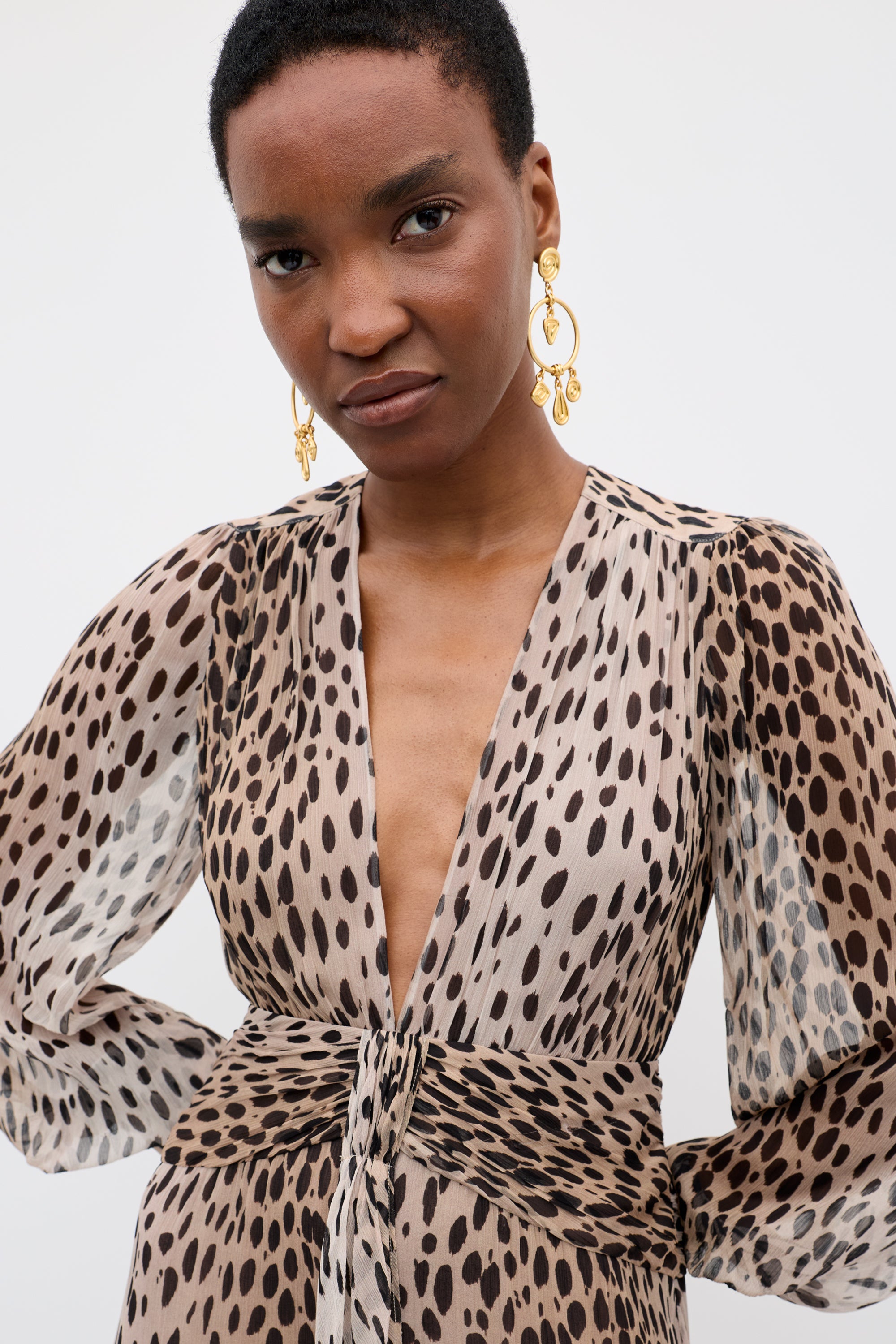 A person with short hair wears gold statement earrings and a long-sleeved Meera - Antelope Spot Black gown in crinkled chiffon with a deep V-neck and animal print, standing against a plain light background.