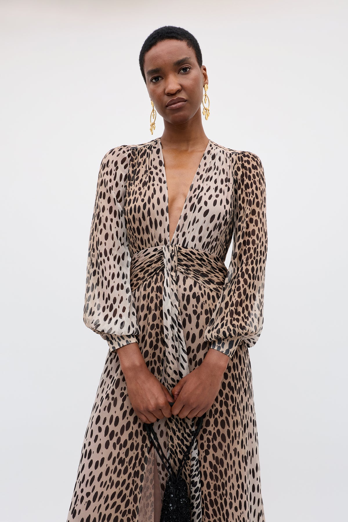 A person stands against a plain background, wearing the Meera - Antelope Spot Black gown—a long-sleeved, deep V-neck dress in tan and black animal print crinkled-chiffon. They have short hair and gold statement earrings.