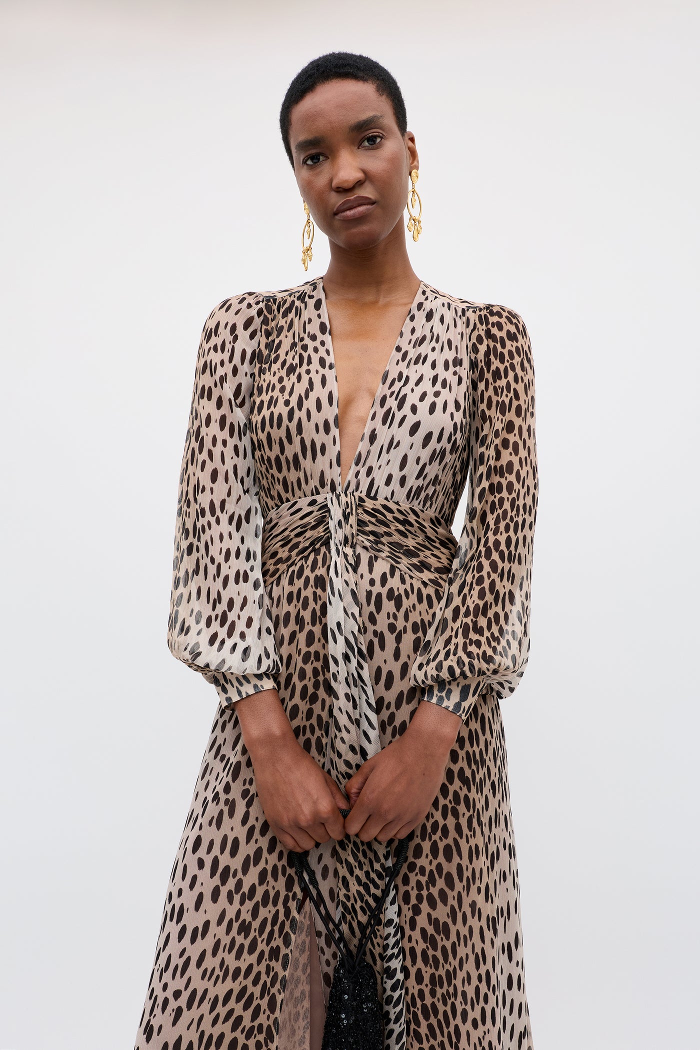 A person stands against a plain background, wearing the Meera - Antelope Spot Black gown—a long-sleeved, deep V-neck dress in tan and black animal print crinkled-chiffon. They have short hair and gold statement earrings.
