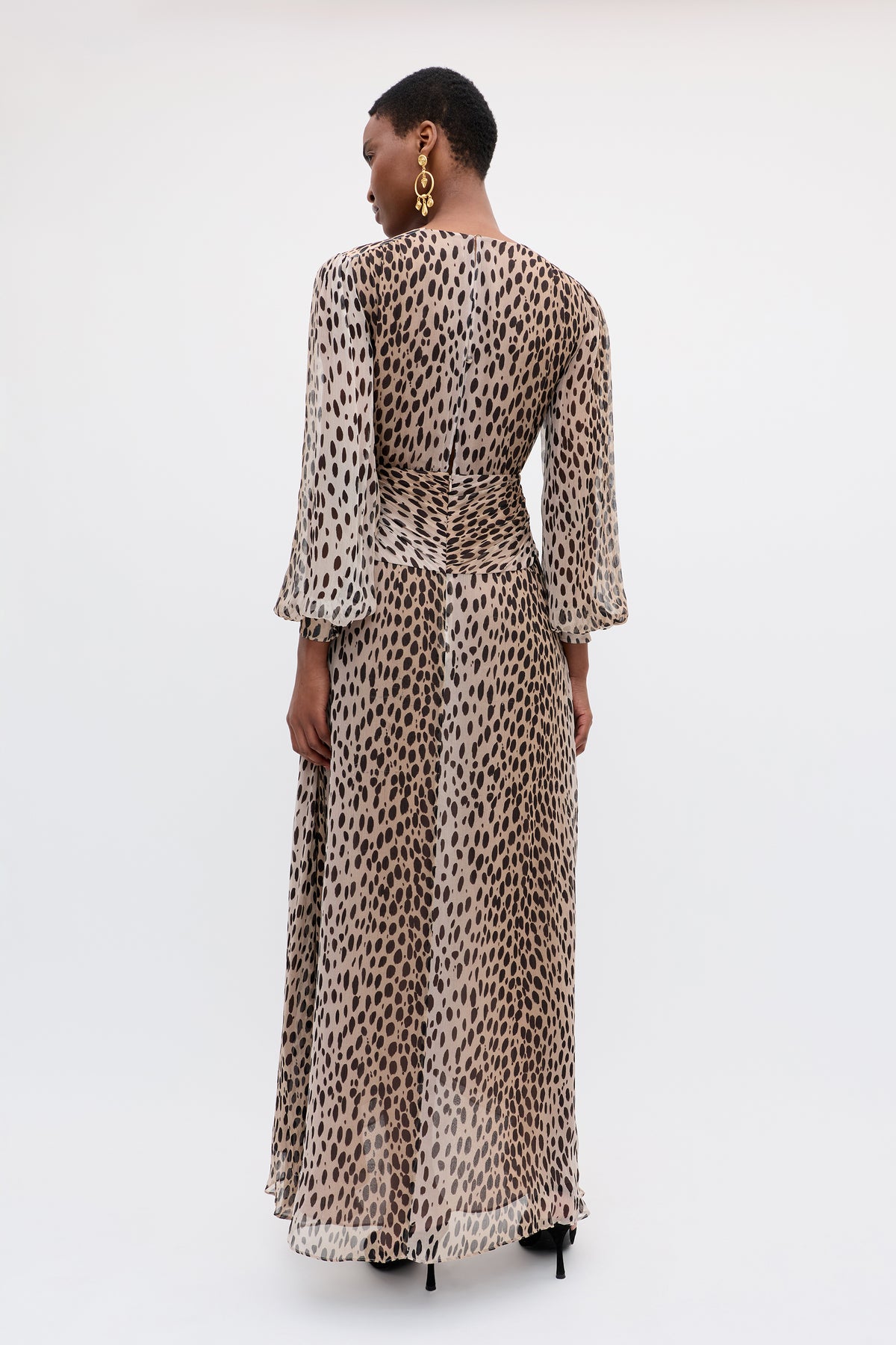 A person in the Meera - Antelope Spot Black gown, a leopard-print crinkled-chiffon dress with long sleeves, stands facing away. With short hair, gold statement earrings, and black heels, they pose against a light background.