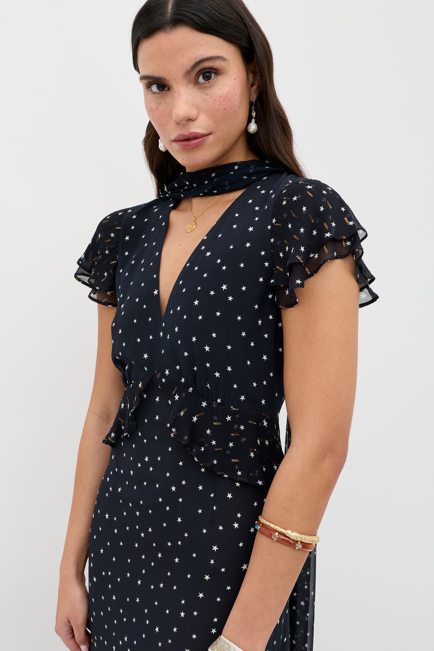 A woman with long dark hair wears the Thanvi - Starry Night Navy dress with georgette ruffles, a deep V-neck, matching scarf, and accessorizes with gold jewelry and pearl earrings against a plain background.