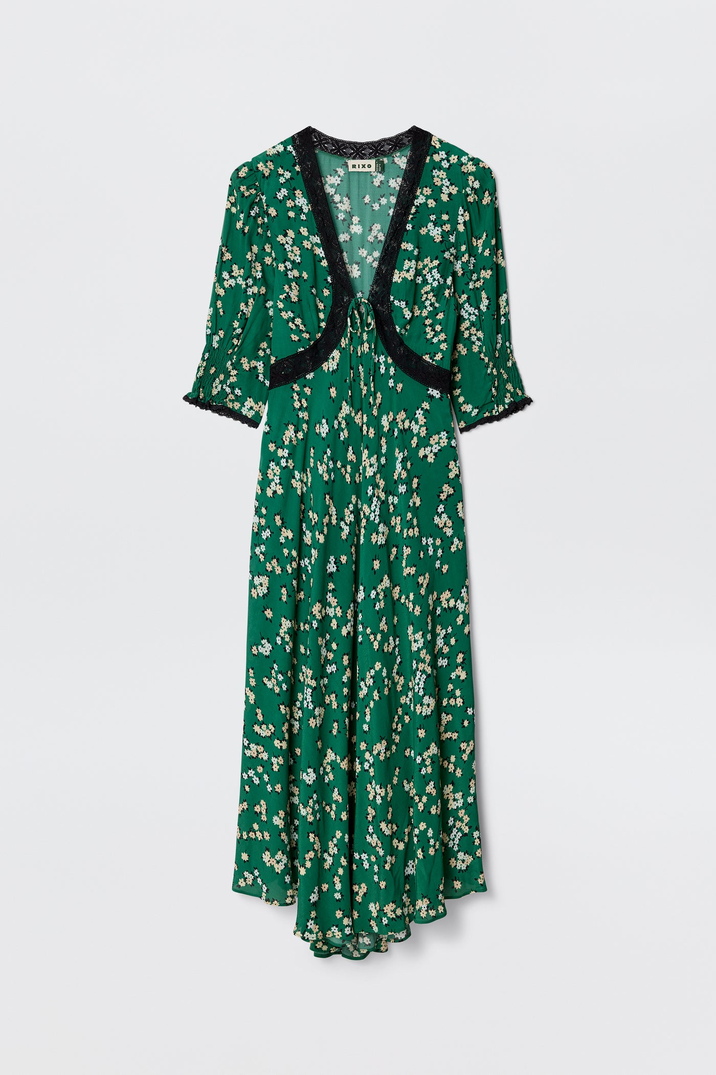 The Amina - City Floral Olive dress features an olive green midi length, short sleeves, and black lace trim at the neckline and cuffs. Its flowy silhouette is adorned with a charming white and yellow daisy print for a vintage tea dress vibe.