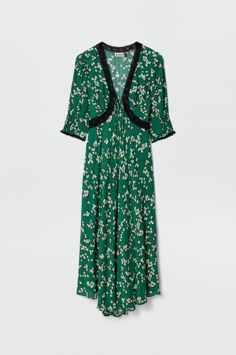 The Amina - City Floral Olive dress features an olive green midi length, short sleeves, and black lace trim at the neckline and cuffs. Its flowy silhouette is adorned with a charming white and yellow daisy print for a vintage tea dress vibe.