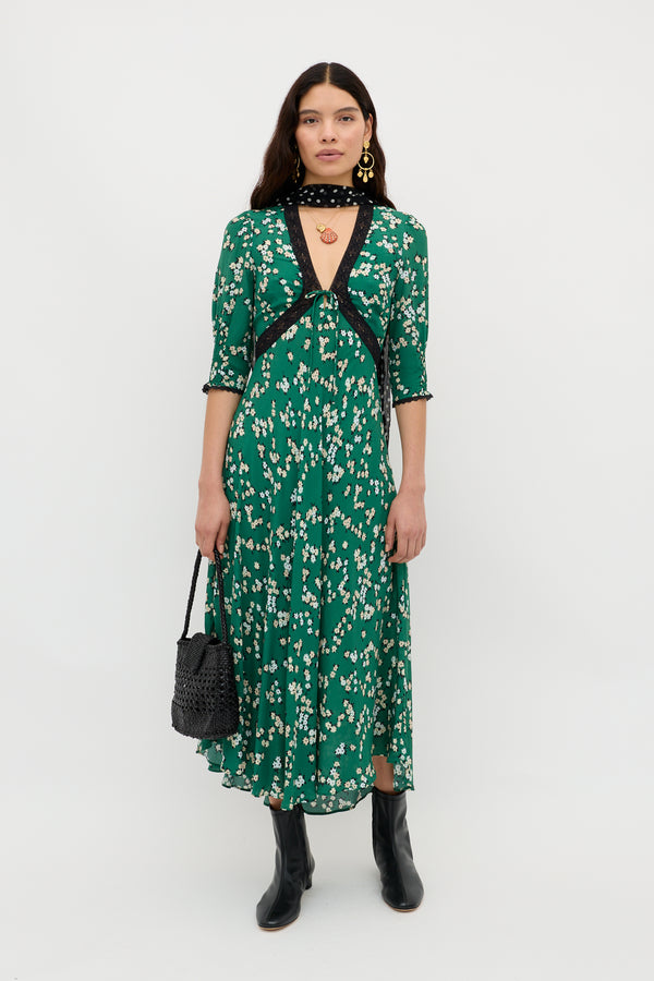 A woman poses against a white background in the Amina - City Floral Olive dress, styled with black lace-trim details, black boots, a neck scarf, gold earrings, and a black woven handbag for a vintage tea dress look.