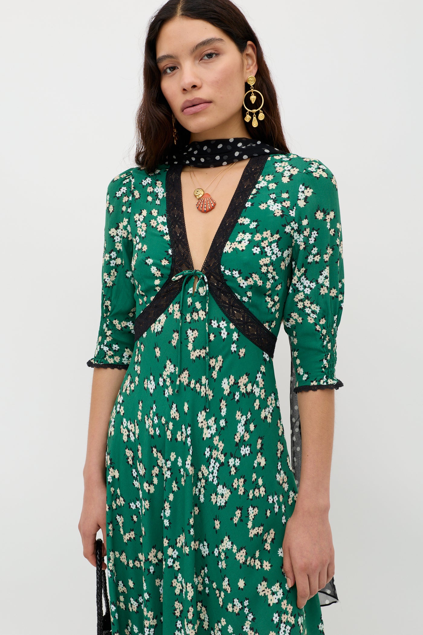 A woman in the Amina - City Floral Olive vintage tea dress with green floral patterns and black lace stands against a plain background, styled with a sheer polka-dot scarf, layered necklaces, and a small black item in her right hand.
