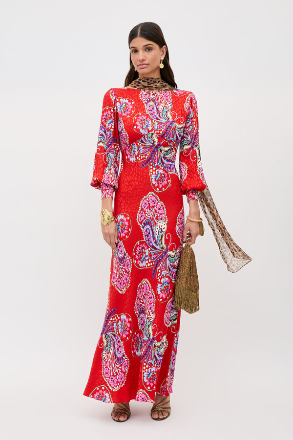 A woman wears the Marni Butterfly Bloom Jacquard Red dress with colorful paisley patterns, gold earrings, bracelets, a fringe handbag, and strappy sandals. She has long dark hair and stands against a white background.
