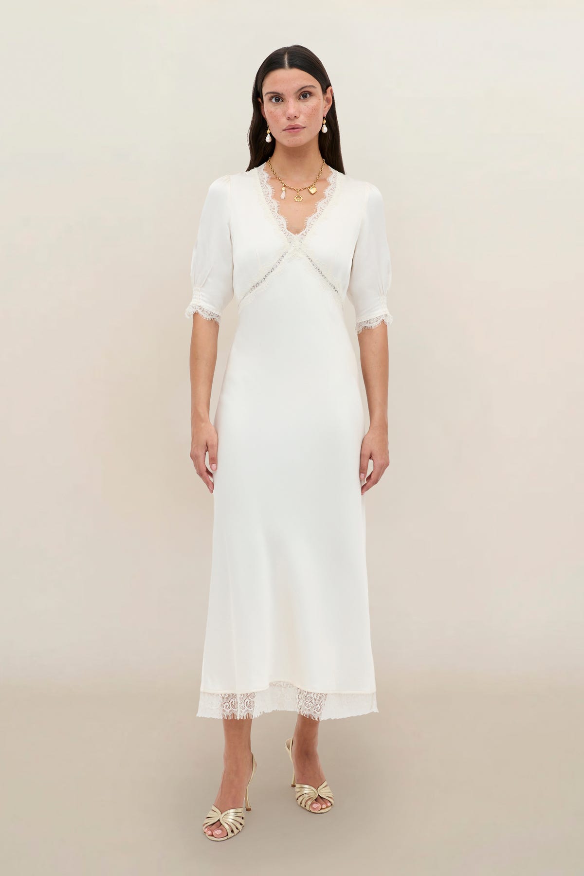 A woman with long dark hair stands against a plain light background, wearing the Annina - Pearl dress—a white, mid-length vintage style with lace trim and puff half sleeves—paired with cream heeled sandals and gold jewelry.