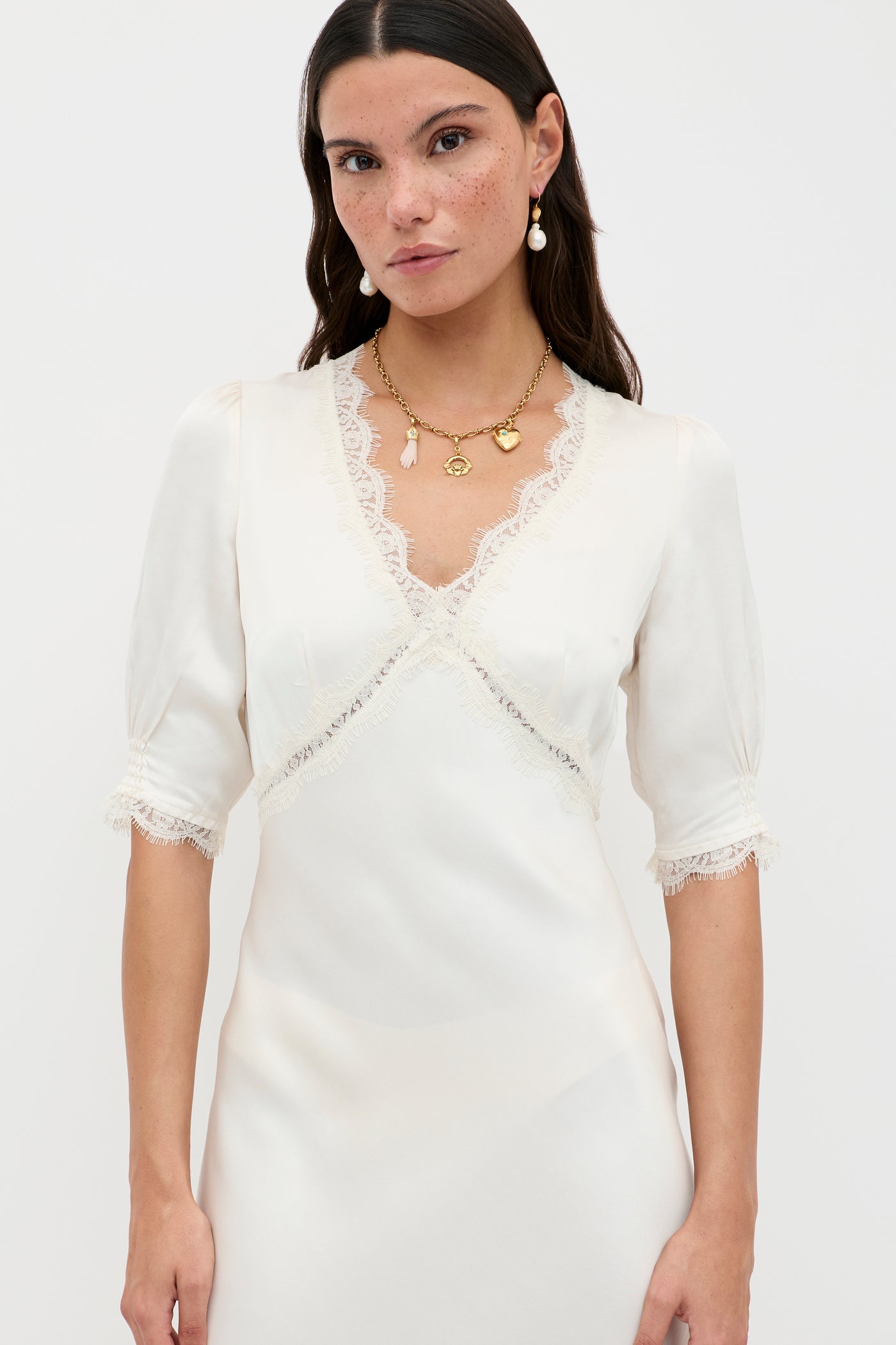 A woman with long brown hair wears the Annina - Pearl, a cream vintage dress featuring lace trim on the V-neckline and sleeves. She pairs it with drop earrings and a gold necklace, standing against a plain white background.