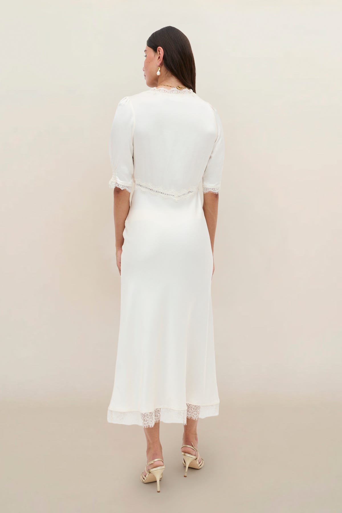 A woman with long dark hair, heeled sandals, and earrings stands facing away against a light background, wearing the Annina - Pearl dress featuring vintage-inspired lace trim and short sleeves.