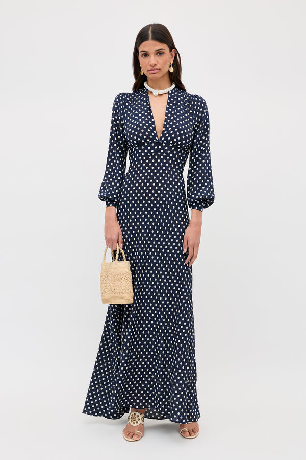 A woman poses against a white background wearing the Emory - Runway Spot Navy dress, featuring long sleeves and a deep V-neckline. She accessorizes with a small woven handbag, sandals, and a statement necklace.