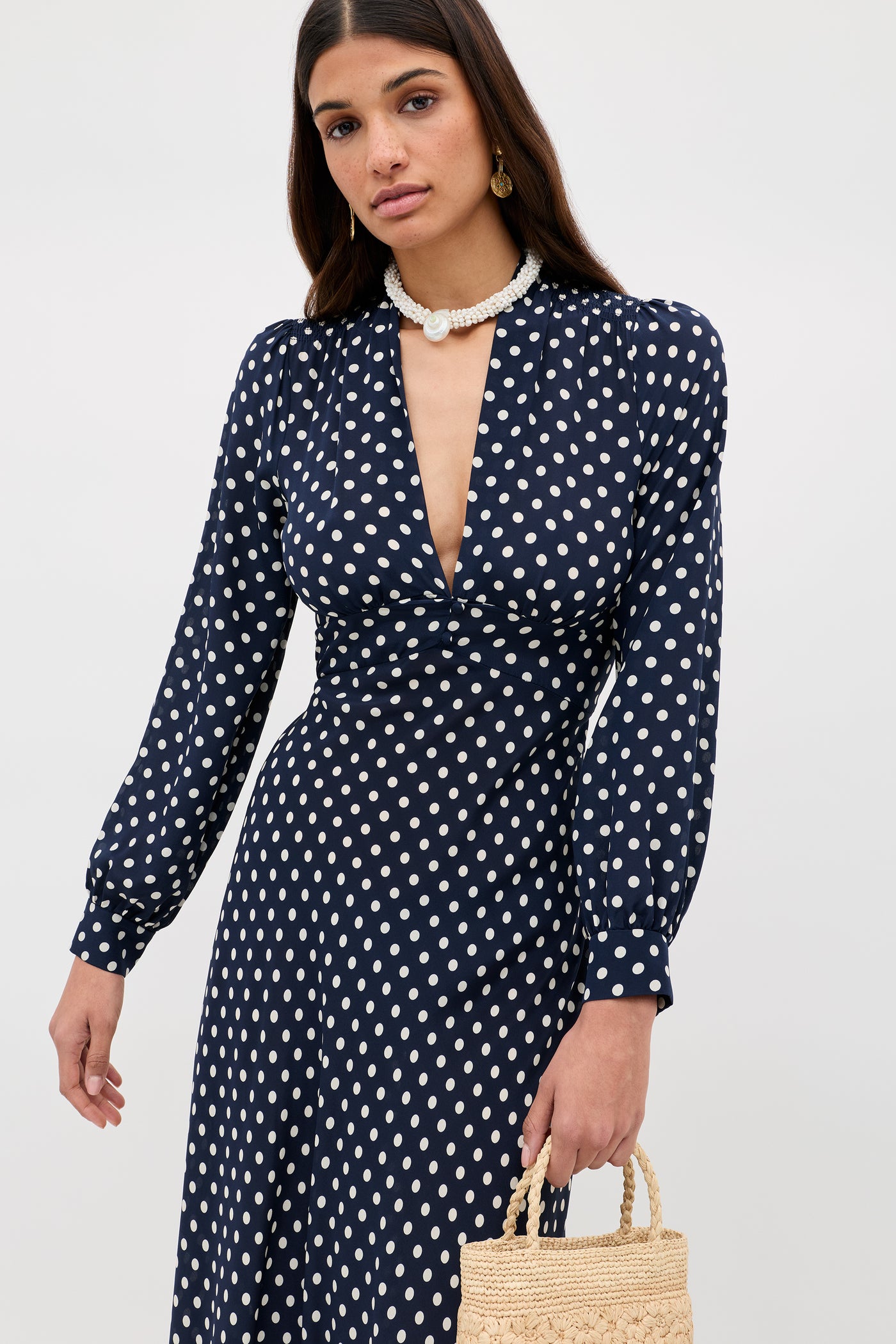 A woman models the Emory - Runway Spot Navy dress, featuring long sleeves and a V-neck, styled with a woven straw handbag and pearl necklace against a plain light background.