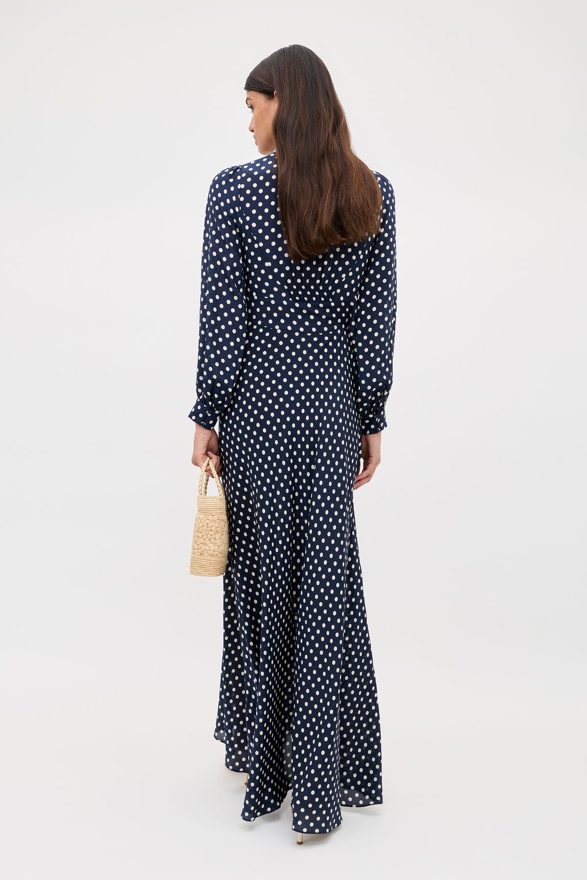 A woman with long brown hair, seen from behind, wears the Emory - Runway Spot Navy dress with white polka dots and holds a small woven straw handbag. The background is plain and light-colored.