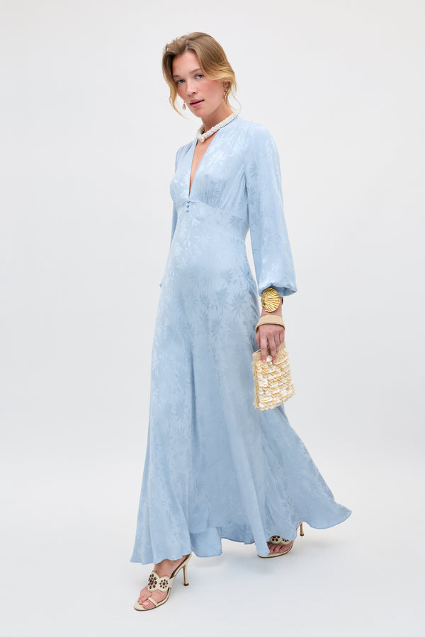 A woman wears the Emory - Daisy Jacquard Bluebell: a light blue, long-sleeved, floor-length dress with a subtle floral pattern. She holds a woven handbag, wears white heels, and stands against a plain background with loosely styled light hair.