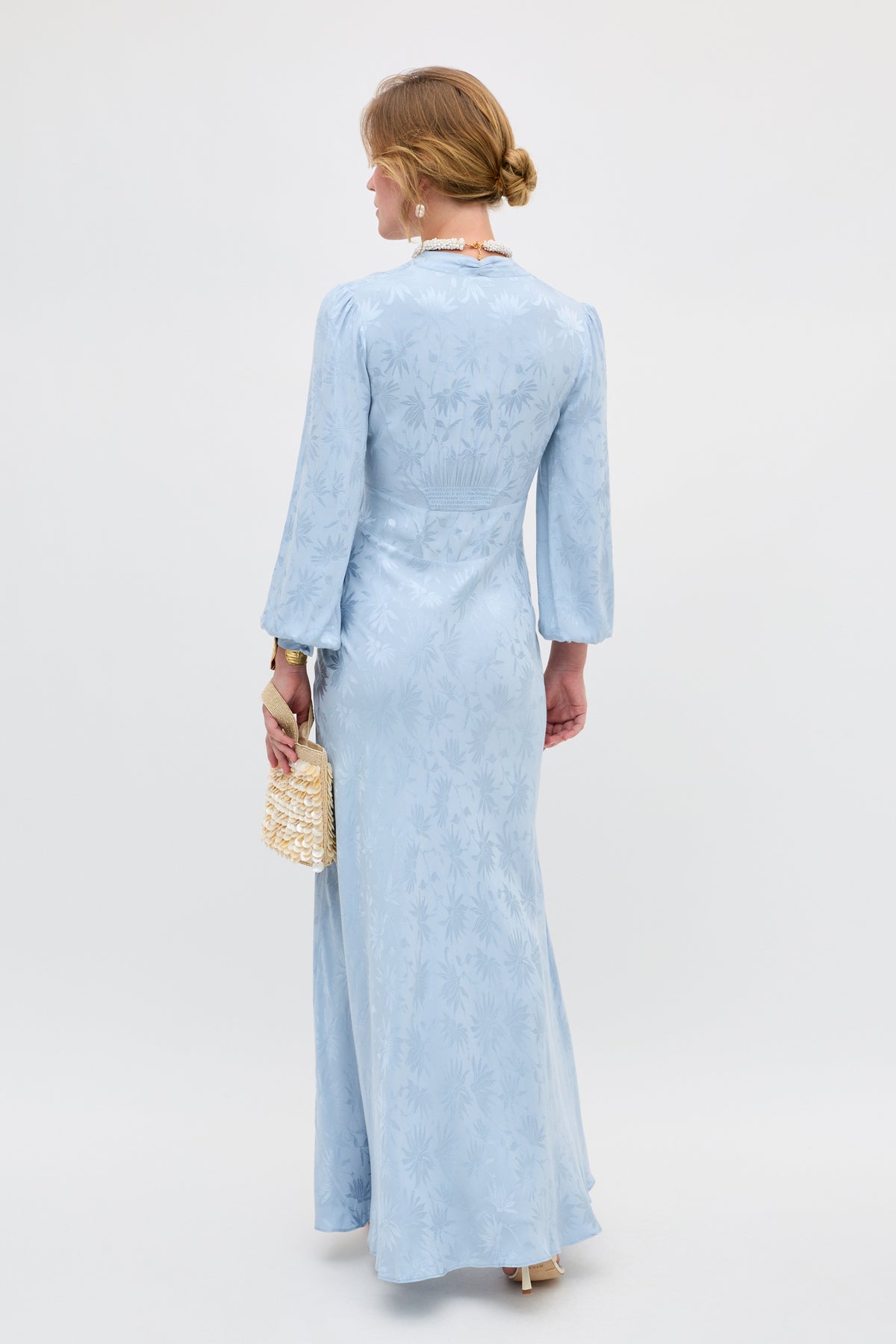 A woman with blonde hair in a low bun wears the Emory - Daisy Jacquard Bluebell, a long, light blue patterned dress with long sleeves. She holds a small woven handbag and stands facing away from the camera against a plain background.