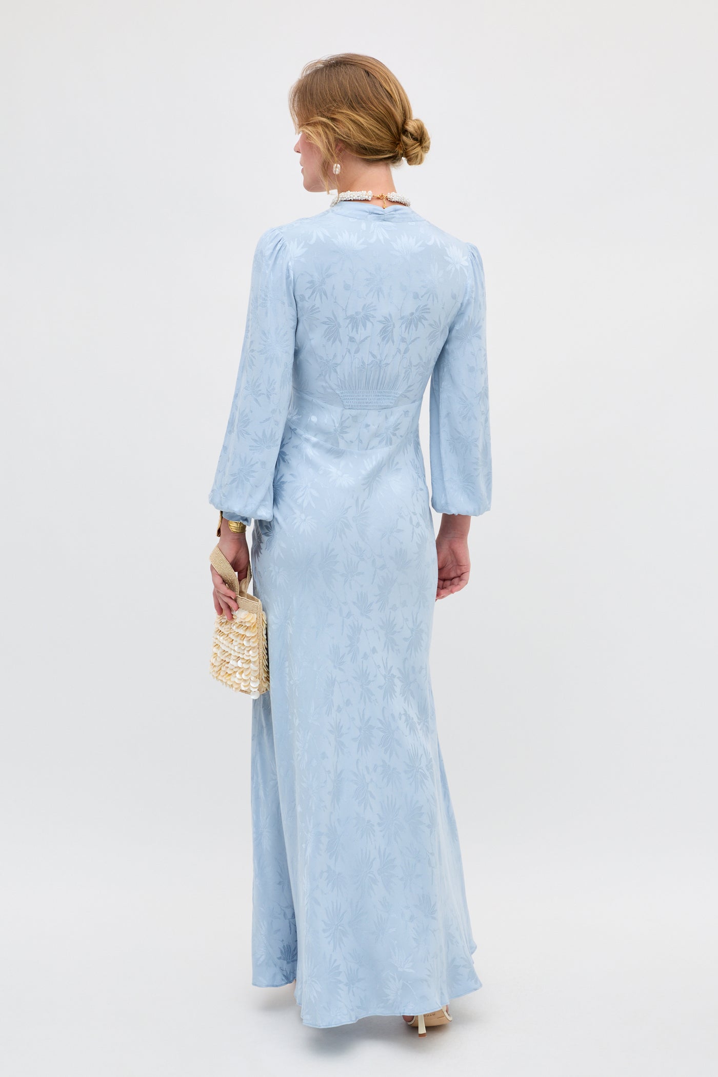 A woman with blonde hair in a low bun wears the Emory - Daisy Jacquard Bluebell, a long, light blue patterned dress with long sleeves. She holds a small woven handbag and stands facing away from the camera against a plain background.