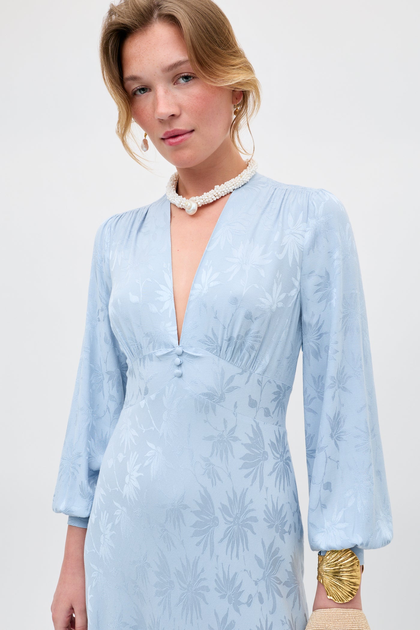A woman wears the Emory - Daisy Jacquard Bluebell, a light blue long-sleeved dress with a subtle floral pattern and deep V-neckline, paired with pearl accessories, standing against a plain background.