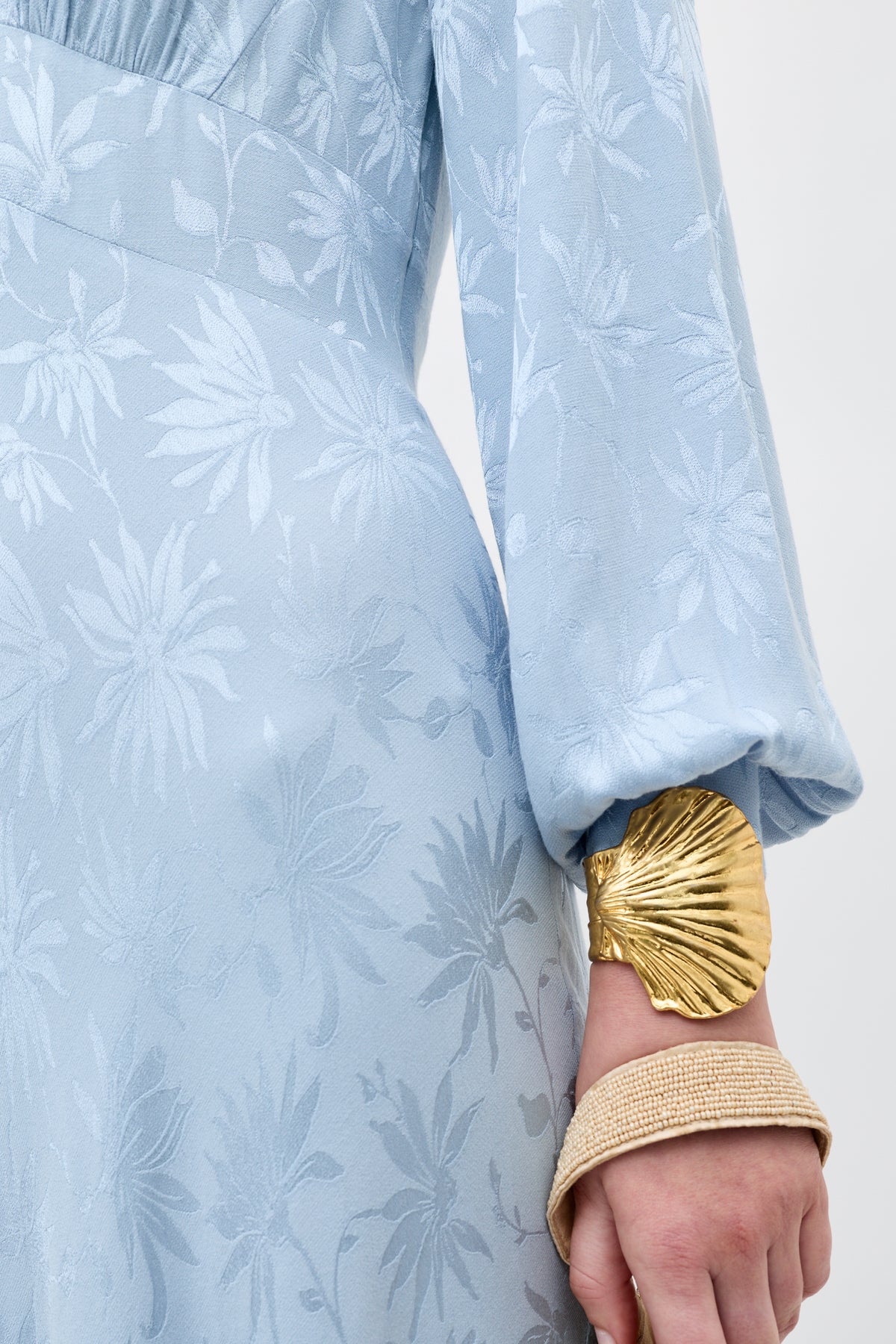 A person wears the Emory - Daisy Jacquard Bluebell dress, featuring light blue long sleeves and a floral pattern, with a large gold shell-shaped bracelet and a textured gold bangle on their wrist.
