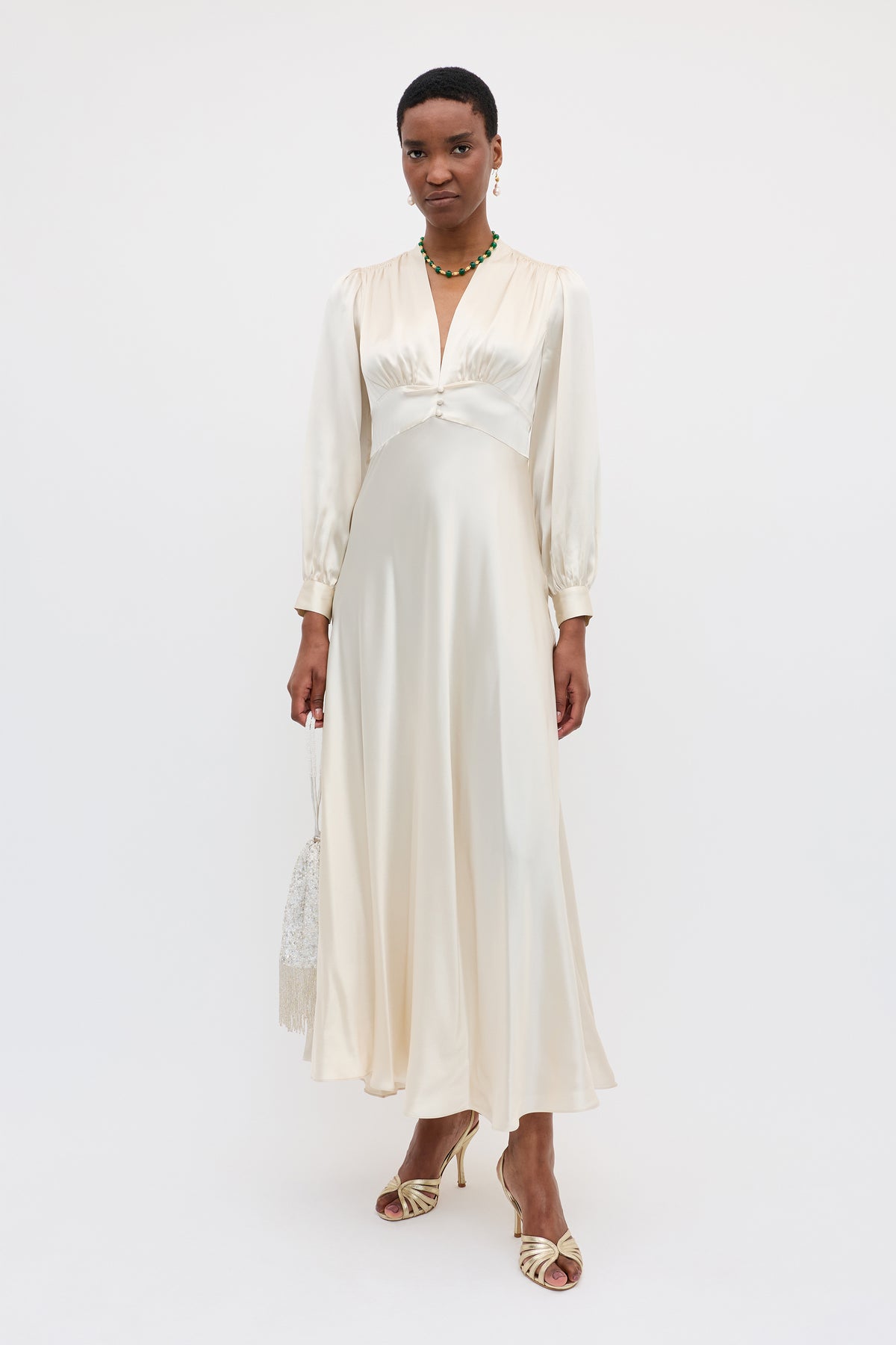 A woman in the Emory - Pearl gown, featuring long sleeves and a deep V-neck, stands against a white background. She accessorizes her silk dress with heeled sandals, a beaded handbag, and a necklace.