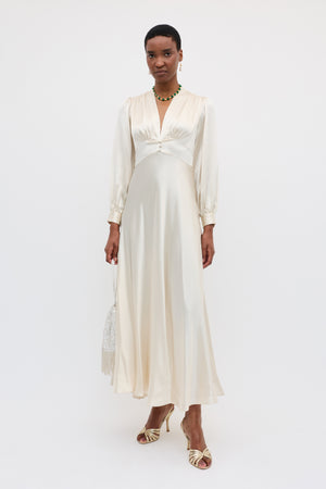 A woman in the Emory - Pearl gown, featuring long sleeves and a deep V-neck, stands against a white background. She accessorizes her silk dress with heeled sandals, a beaded handbag, and a necklace.