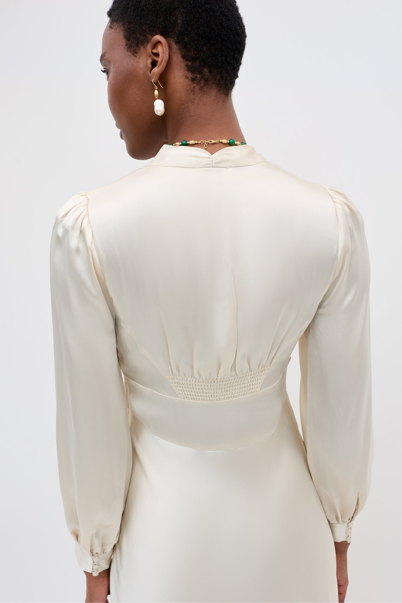 A person with short hair and pearl earrings is shown from the back in the Emory - Pearl gown, highlighting its gathered waist and gently puffed shoulders in a soft pearl tone.