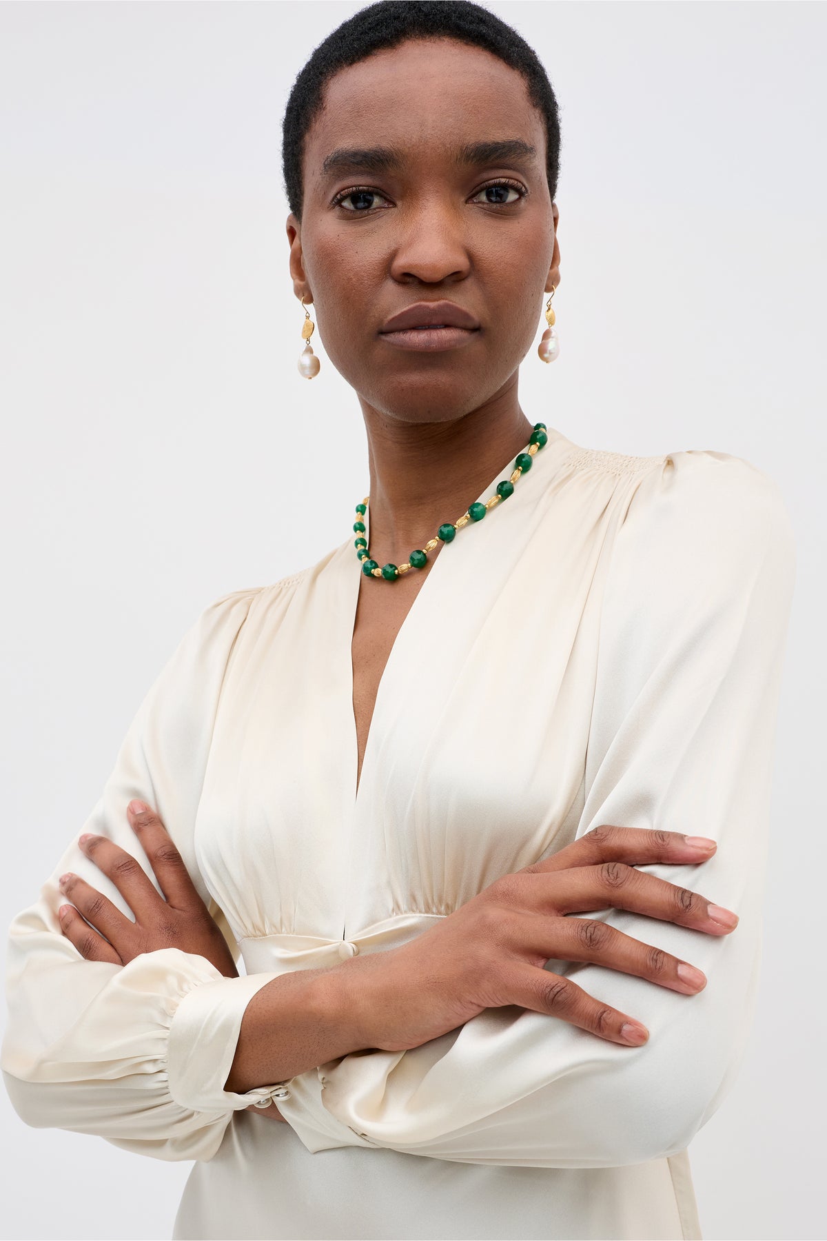 A woman with short hair wears a cream long-sleeve blouse, green beaded necklace, and drop earrings. She stands confidently with arms crossed against a light backdrop inspired by the Emory - Pearl colourway from RIXO.