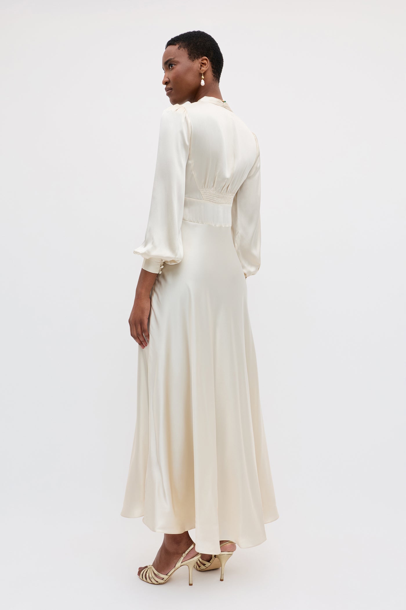 A woman in the Emory - Pearl gown with long sleeves and high heels stands against a plain white background, looking over her shoulder.