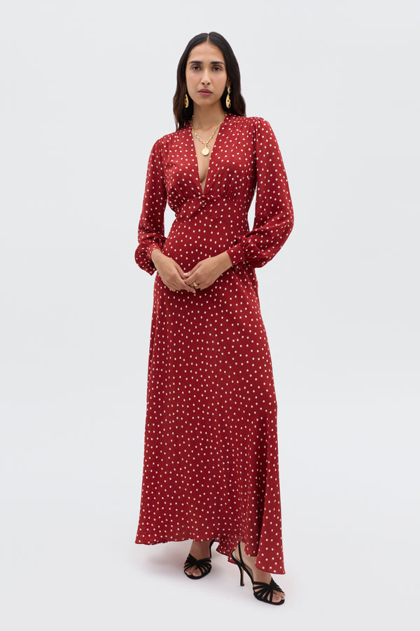 A woman poses against a plain background wearing the Emory - Layla Dot Red dress, featuring long sleeves, a deep V-neckline, and a red polka dot pattern. She pairs it with black high heels, layered gold necklaces, and flowing dark hair.