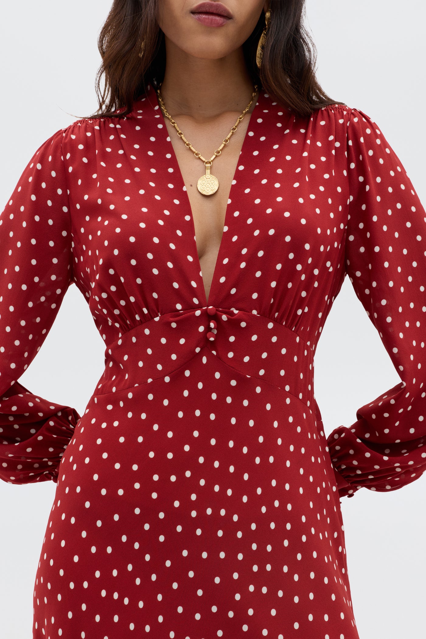 A woman wears the RIXO Emory - Layla Dot Red dress, featuring red polka dots, long sleeves, and a deep V-neckline. She has wavy dark hair and accessorizes with a gold pendant necklace and gold earrings.