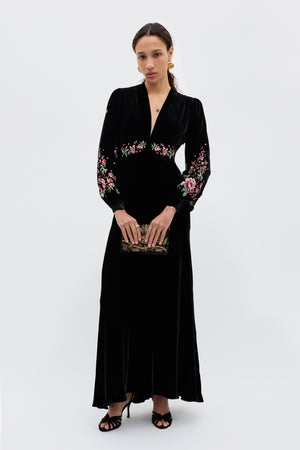 A woman stands against a plain background wearing the Emory - Blossom Embroidery Pink dress, featuring 1930s-inspired floral detailing on the sleeves and waist. She accessorizes with a small clutch, black heeled sandals, and gold earrings.