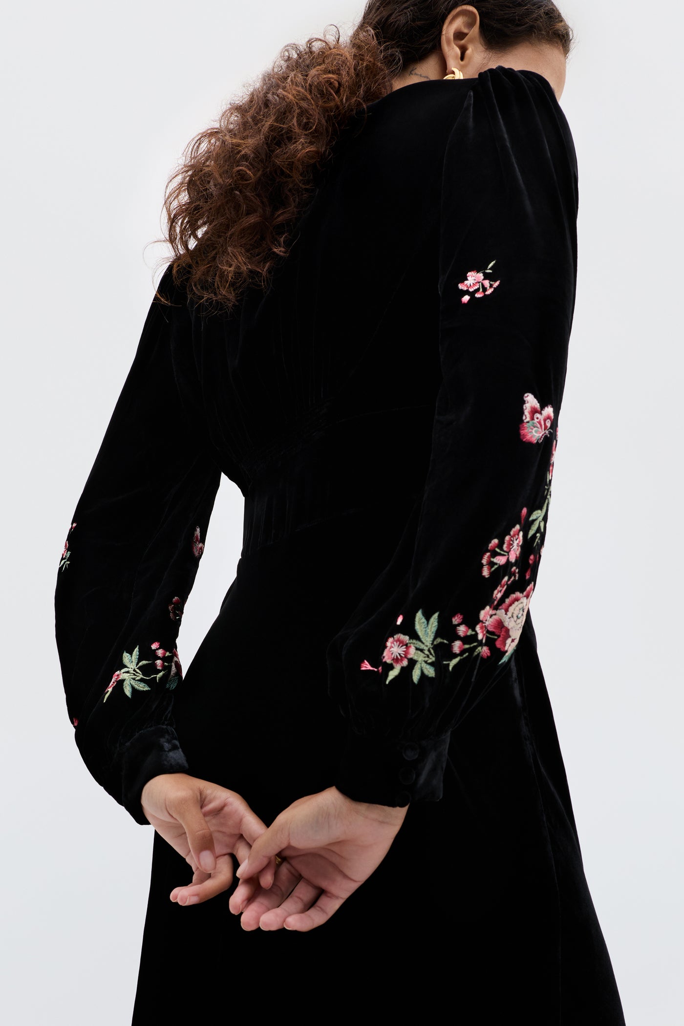 A person with curly brown hair stands with their back to the camera, hands clasped, wearing the Emory - Blossom Embroidery Pink dress featuring 1930s-inspired colorful floral embroidery on the sleeves.